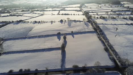 Cold-snowy-winter-British-patchwork-farmland-countryside-rural-scene-aerial-slow-forward-shot-at-sunrise