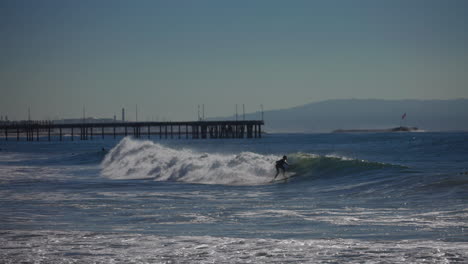 A-surfer-rides-a-wave-down-the-line-with-a-pier-and-mountains-in-the-background