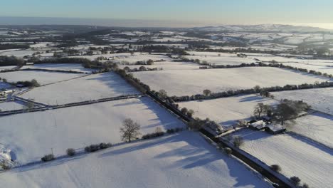 Cold-snowy-winter-British-patchwork-farmland-countryside-rural-scene-aerial-at-sunrise-cinematic-left-orbit