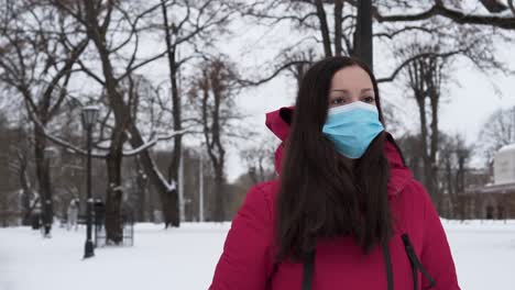 Weibliche-Touristin,-Die-Kapuzenmantel-Mit-Gesichtsmaske-Trägt,-Die-Im-Verschneiten-Winterpark-Spazieren-Geht