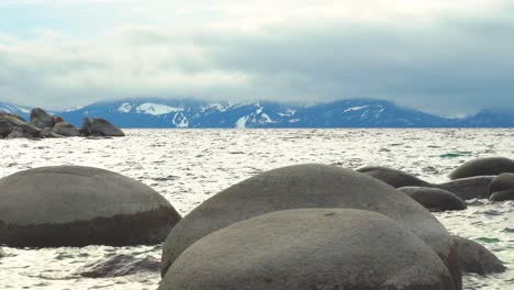 Boulders-sticking-out-of-the-water-in-Lake-Tahoe,-Nevada-with-snow-capped-mountains-in-the-background-during-a-cloudy-foggy-day---Sand-Harbor
