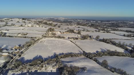 Cold-snowy-winter-British-patchwork-farmland-countryside-rural-scene-aerial-at-sunrise-slow-cinematic-pan-right