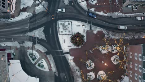 Top-down-aerial-of-traffic-passing-through-urban-city-street-with-snow-and-Christmas-tree,-lights