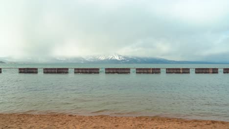 South-Lake-Tahoe-during-winter-with-a-sandy-golden-beach-foreground,-pier-in-the-middle-and-gorgeous-snow-capped-mountains-in-the-background---wide-shot
