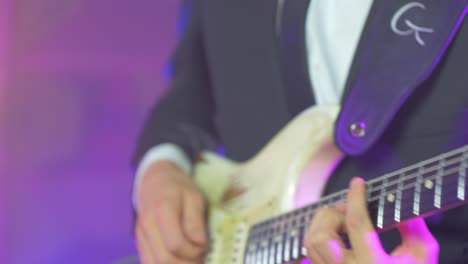 Close-up-of-a-professional-musician-in-a-black-suit-playing-chords-on-an-electric-stratocaster-guitar-during-a-live-session-on-stage-with-warm-studio-lights-in-the-blurred-background