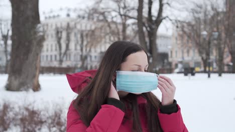 Caucasian-Woman-In-Red-Hooded-Coat-Puts-On-Medical-Mask-In-Winter-Park