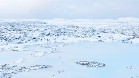 Family-walking-on-snow-covered-shore-of-blue-lagoon-in-Iceland,-aerial