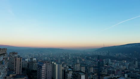 Man-Smoking-On-The-Rooftop-Overlooking-Tbilisi-Cityscape-In-Georgia-With-Contrail-In-The-Sky-On-A-Sunrise,-aerial-drone