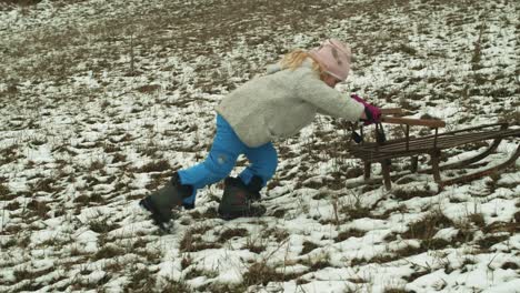 Determined-girl-pushes-sledge-up-a-hill-with-lack-of-snow,-slow-motion