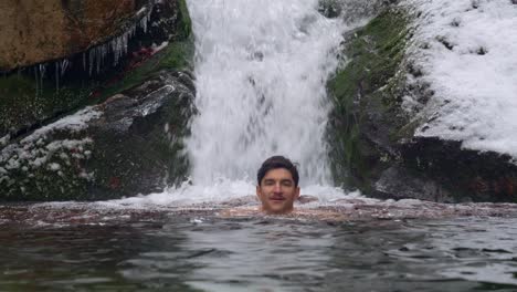 Adult-male-ice-swimming-in-icy-cold-water-under-a-picturesque-waterfall-in-mountains-during-winter-day---static-shot,-slow-motion
