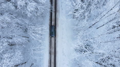 Top-Down-View-of-a-car-driving-through-a-snow-covered-Forest-Road,-Aerial-drone-hovering-over-village-road-in-winter-when-a-car-passes-by