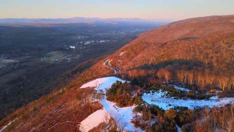 Flying-high-over-a-mountain-with-a-light-snow-covering,-and-scenic-road-with-a-large,-expansive-valley-below-with-mountains-in-the-distance-during-sunset