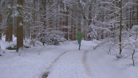 Athlethic-adult-man-running-from-distance-in-green-outfit-in-snow-covered-forest-on-wild-path-during-cold,-winter-day---toward-camera,-slow-motion
