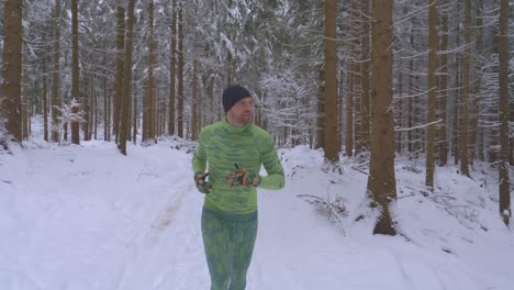 Athletic-man-in-green-outfit-running-in-woods-on-snowy-trail-during-cold-winter-day---follow-shot,-slow-motion
