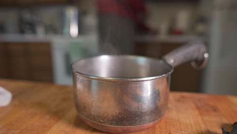 Steam-Rising-From-Stainless-Pot-Place-On-Wooden-Table-With-Blurry-Image-Of-Man-At-Kitchen