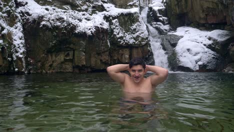 Handsome-man-standing-in-subzero-cold-water-by-the-beautiful-waterfall-in-snowy-mountains-during-winter-day---static-shot,-slow-motion