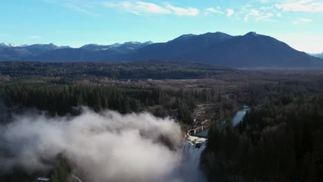 Aerial-backwards-shot-of-beautiful-snoqualmie-falls-hiding-by-fog-blanket-and-clear-wide-shot-of-forest-and-mountains-in-backdrop