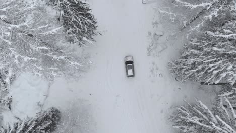 Aerial-view-overlooking-a-slow-racing-car-on-a-road,-in-middle-of-snowy-forest,-winter,-cloudy-day---drone-shot,-top-down,-tracking