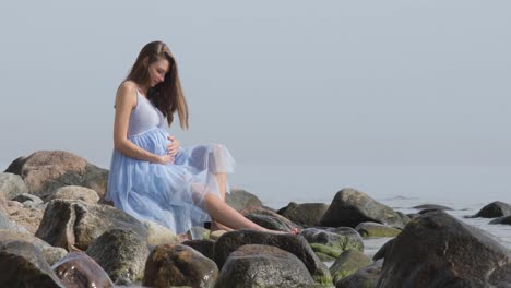 A-pregnant-woman-sits-on-the-rocks,-deep-in-thought-as-she-holds-her-belly-and-thinks-about-her-babies-future