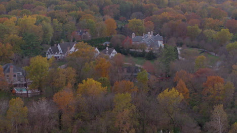 Aerial-view-of-beautiful-large-houses-nestled-in-trees-at-peak-color-in-Fall-with-a-tilt-up-to-a-pretty-pink-and-yellow-hombre-sky-at-sunset