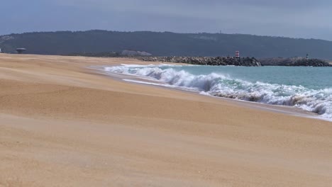 Zeitlupenwellen-Weiß-Mit-Schaum-Bedecken-Den-Nassen-Sand-Des-Strandes,-Klippe-Mit-Leuchtturm-Im-Hintergrund