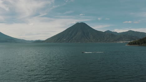 Vista-Aérea-De-Drones-De-Un-Barco-Que-Viaja-En-El-Lago-Atitlán,-Guatemala-Con-Hermoso-Volcán-Y-Paisaje-De-Agua-Azul