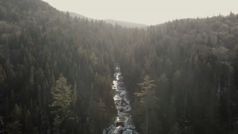 Stream-Flowing-Gently-Through-The-Icy-Rock-Path-Down-From-The-Lush-Forest-Mountain-In-Vallee-Bras-du-Nord,-Quebec,-Canada---Aerial-Drone-Shot
