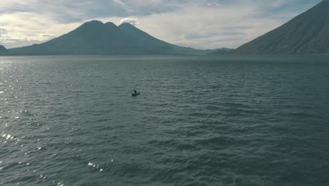 Drone-aerial-shot-of-lake-Atitlan,-Volcanoes-and-a-local-man-kayaking-in-Guatemala