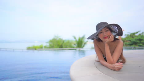 Mujer-Sonriente-Asiática-Con-Sombrero-Tomando-El-Sol-Tumbado-En-El-Borde-De-La-Piscina