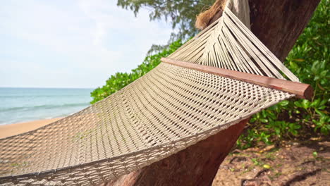 Close-up-of-an-empty-woven-hammock-rocking-in-the-breeze,-waves-rolling-in-on-a-sandy-beach-in-the-background