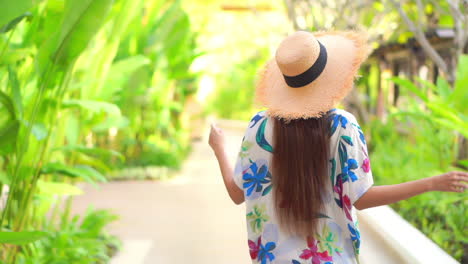 Back-of-stylish-young-female-in-colorful-summer-dress-and-hat-walking-on-pathway-with-tropical-plants-on-sunny-day,-slow-motion