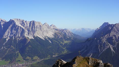 Vista-De-La-Cruz-En-La-Cumbre-De-Grubigstein-Con-Dos-Excursionistas-Disfrutando-De-Las-Increíbles-Vistas-De-Zugspitze,-Ehrwald-Y-Lermoos-En-Las-Montañas-De-Los-Alpes