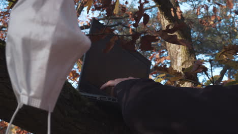Hands-typing-laptop-in-forest-during-covid-pandemic,-face-mask-hanging-on-tree,-closeup-shot