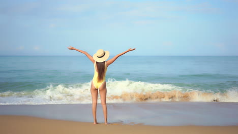 A-young-woman-in-a-one-piece-bathing-suit-with-her-back-to-the-camera-greets-the-incoming-ocean-tide-and-waves-with-joy-and-extended-arms