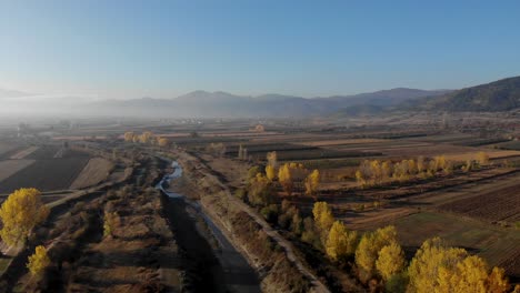 River-and-brown-field-with-yellow-trees-on-mountain-background-covered-in-fog