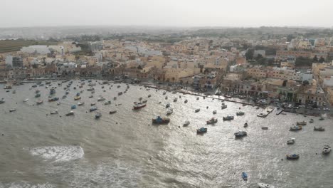 Aerial-Wide-Pan-Right-Shot-Of-Marsaxlokk-Port-With-Village-Background-In-Malta