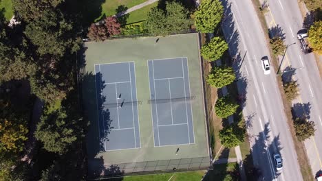 2-2-Aerial-hold-top-birds-eye-view-of-couples-playing-tennis-at-a-modern-park-by-luxury-club-close-to-a-garden-road-freeway-with-seaguls-and-pigeons-flying-over-the-athletic-adults-meeting-at-the-net