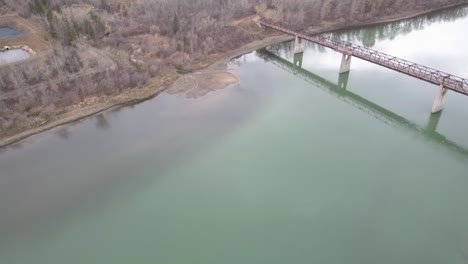 1-3-aerial-flight-over-dreary-river-park-by-a-truss-pedestrian-multimaterial-bridge-of-concrete-wood-and-steel-on-a-fall-afternoon-where-merky-poluted-waters-appear-oil-spilled-clumps-of-toilet-waste