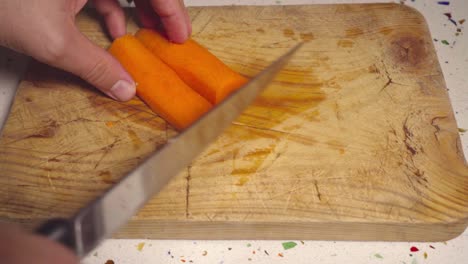 Man-slices-carrot-with-knife-on-wooden-cutting-board,-Closeup-on-Hands