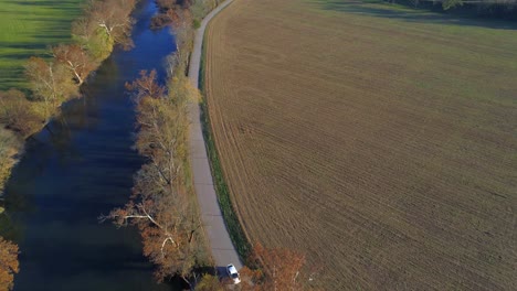 Aerial-shot-tracking-vehicle-traveling-down-a-country-road-through-farm-land-next-to-a-creek