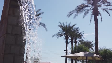 Slow-motion-tilt-down-from-the-spout-of-a-freestanding-water-feature,-a-brick-fountain-surrounded-by-palm-trees-and-city-streets,-Scottsdale,-Arizona