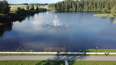 Man-Sitting-Beside-Teperis,-Beautiful-Lake-With-Fountain-In-An-Old-Park-In-Smiltene---ascending-drone,-pullback