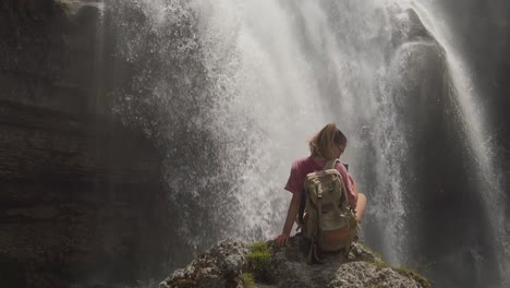 A-girl-hiking-is-sitting-on-a-rock-under-a-waterfall-in-a-nature-reserve