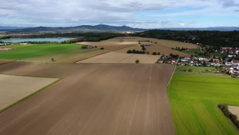 Drone-flight-over-harvested-fields-in-Kaiserstuhl-on-a-cloudy-summerday,-mountains-and-quarry-pond-in-the-background