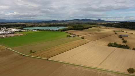 Drone-flight-backwards-from-a-quarry-pond-and-gravel-plant-over-fields-on-a-cloudy-summer-day,-mountains-in-the-background