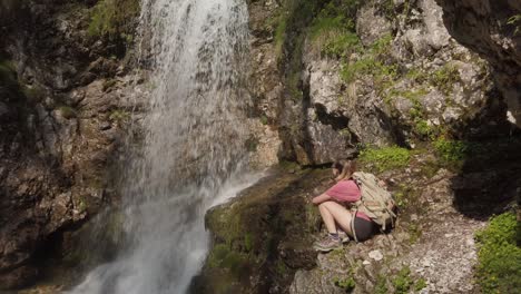 A-girl-hiking-is-sitting-on-a-rock-near-a-waterfall-in-a-nature-reserve