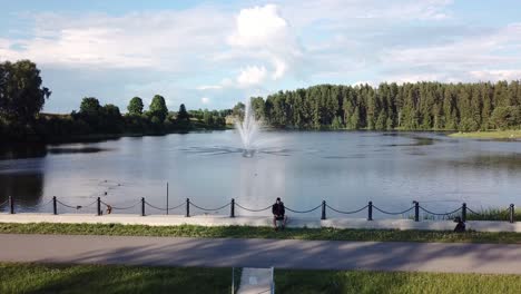 Joven-Sentado-Y-Controlando-Drones-Con-Fuente-En-El-Lago-Teperis-En-Segundo-Plano-Durante-El-Verano-En-Smiltene,-Letonia