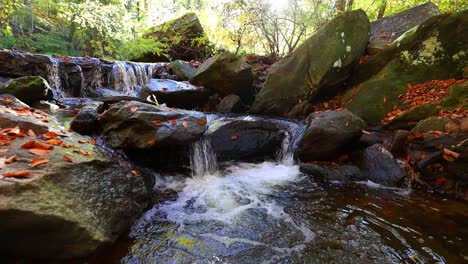 Beautiful-waterfall-cascade-over-large-wet-boulders-with-fallen-leaves-of-Autumn-on-rocks-along-creek-in-park