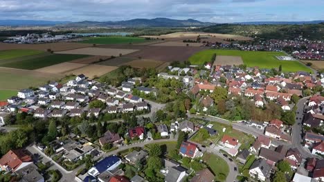 Drohnenflug-über-Das-Dorf-Oberrimsingen-An-Einem-Bewölkten-Sommertag,-Felder,-Berge-Und-Ein-See-Im-Hintergrund
