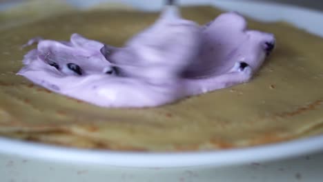 Close-up-view-of-purple-blueberry-cream-being-spread-on-a-french-pancake-on-a-white-plate